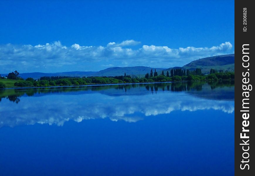 Beautiful blue sky with a few clouds, trees und hill mirrored in calm lake surface. Beautiful blue sky with a few clouds, trees und hill mirrored in calm lake surface