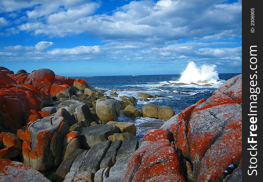 Wave splashing against rocks at coast, with boulders overgrown with orange lichen in foreground and blue sky above