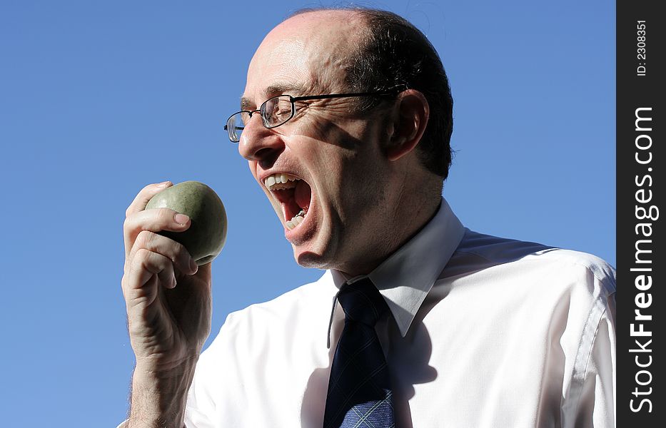 Businessman wearing business clothes about to take a bite into a green apple, showing face, hand, apple and blue sky behind him. Businessman wearing business clothes about to take a bite into a green apple, showing face, hand, apple and blue sky behind him.