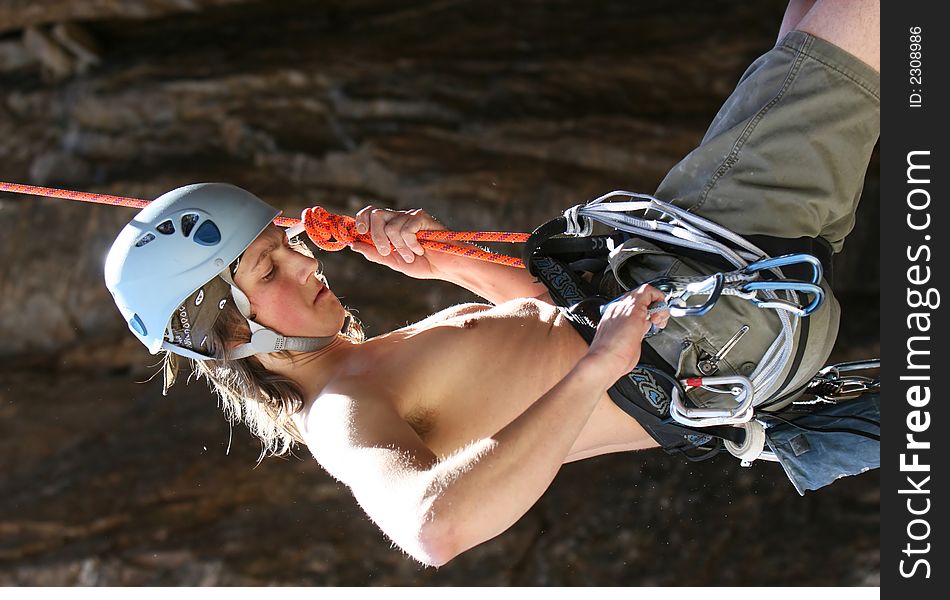 A teenager is lowered off a rock face after completing a climb. He is wearing a harness and helmet. Quickdraws and caribiners hang from his harness. A teenager is lowered off a rock face after completing a climb. He is wearing a harness and helmet. Quickdraws and caribiners hang from his harness.