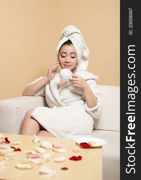 Portrait of pretty Chinese businesswoman in white bathrobe with towel on head speaking on phone with cup of coffee. Portrait of pretty Chinese businesswoman in white bathrobe with towel on head speaking on phone with cup of coffee
