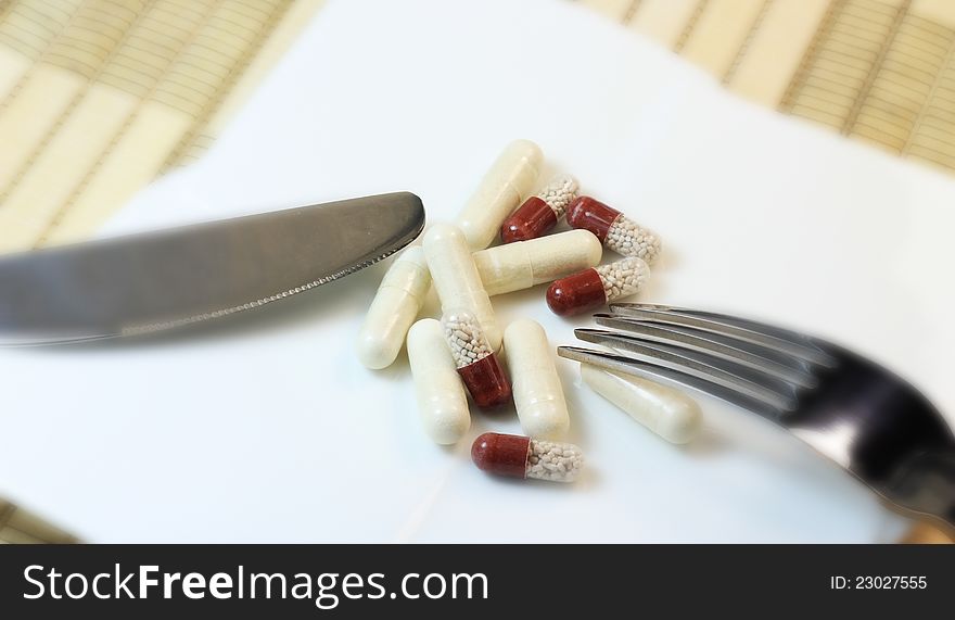 Pills on white plate with knife and a fork, all on table as a concept of taking too much medicine. Shallow DOF.