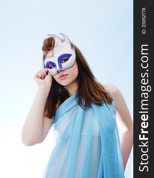 Masquerade Mask held to face of pretty brunette woman. Soft Blue costume and background. Masquerade Mask held to face of pretty brunette woman. Soft Blue costume and background
