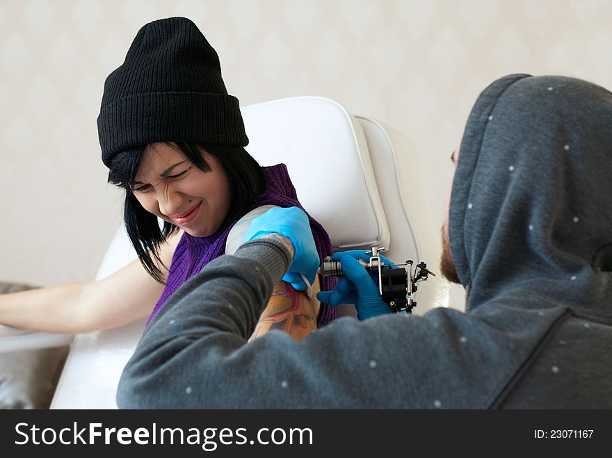 Emotions of a girl while a tattoo artist applying his craft on her shoulder at his workshop. Focus on female's face. Emotions of a girl while a tattoo artist applying his craft on her shoulder at his workshop. Focus on female's face.