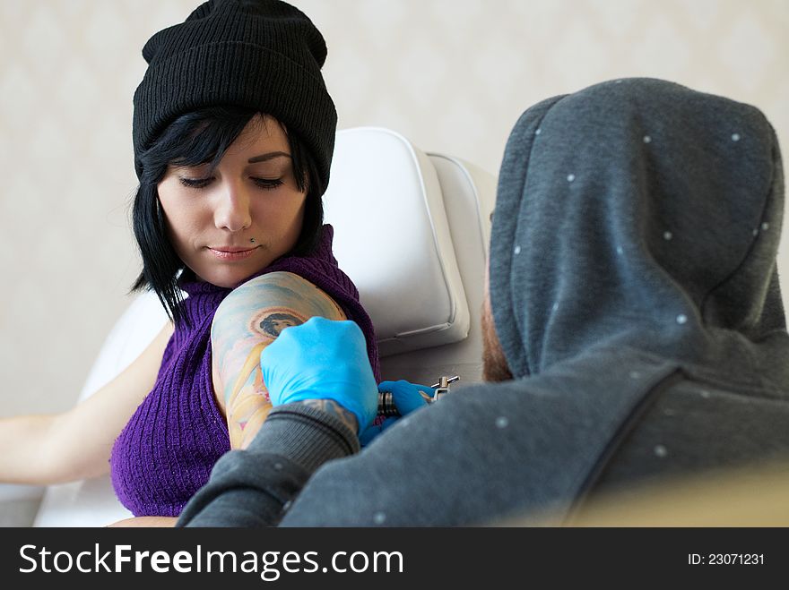 Emotions of a girl while a tattoo artist applying his craft on her shoulder at his workshop. Focus on female's face. Emotions of a girl while a tattoo artist applying his craft on her shoulder at his workshop. Focus on female's face.