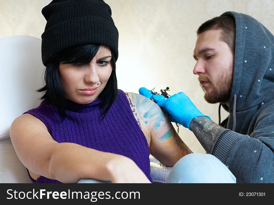 Emotions of a girl while a tattoo artist applying his craft on her shoulder at his workshop. Focus on female's face. Emotions of a girl while a tattoo artist applying his craft on her shoulder at his workshop. Focus on female's face.