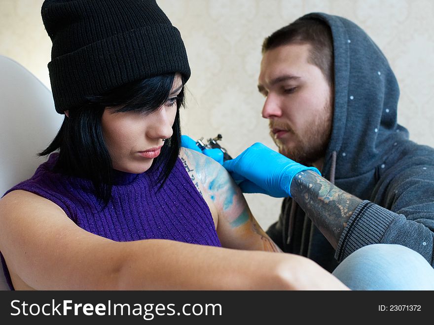 Emotions of a girl while a tattoo artist applying his craft on her shoulder at his workshop. Focus on female's face. Emotions of a girl while a tattoo artist applying his craft on her shoulder at his workshop. Focus on female's face.