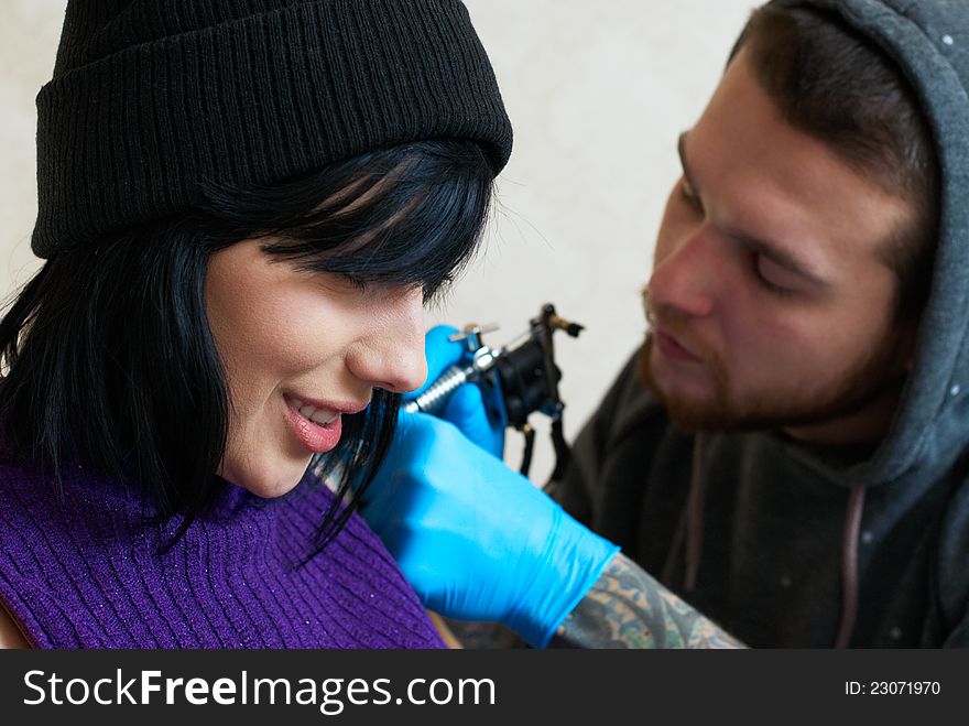 Emotions of a girl while a tattoo artist applying his craft on her shoulder at his workshop. Focus on female's face. Emotions of a girl while a tattoo artist applying his craft on her shoulder at his workshop. Focus on female's face.