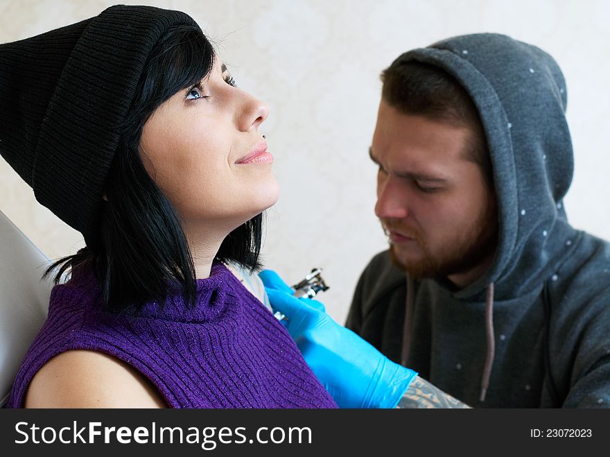Emotions of a girl while a tattoo artist applying his craft on her shoulder at his workshop. Focus on female's face. Emotions of a girl while a tattoo artist applying his craft on her shoulder at his workshop. Focus on female's face.