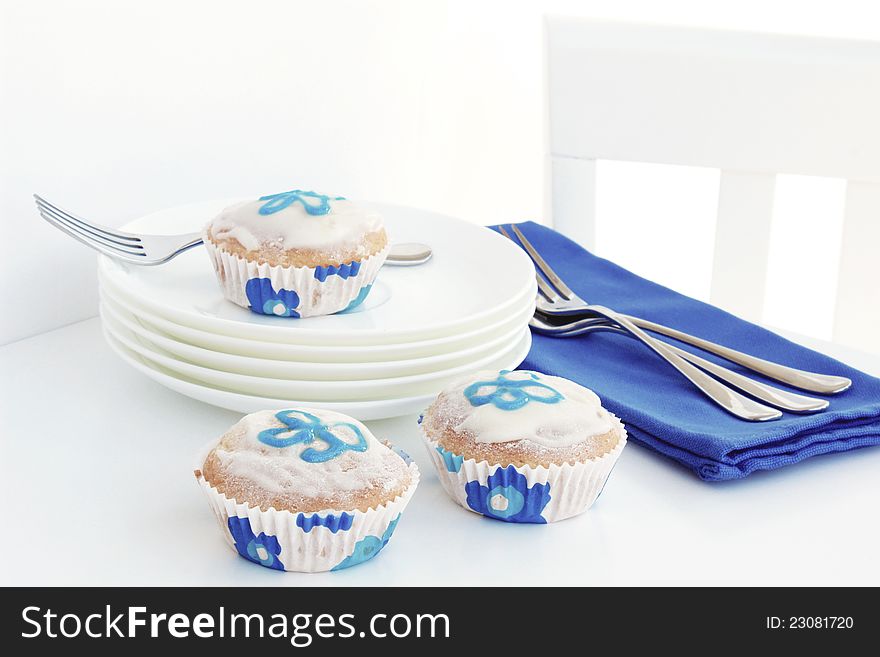 A high key image of blue and white coloured cupcakes with a flower on the icing, surrounded by blue serviettes, white plates and stainless steel forks on a white table a chair. A high key image of blue and white coloured cupcakes with a flower on the icing, surrounded by blue serviettes, white plates and stainless steel forks on a white table a chair.