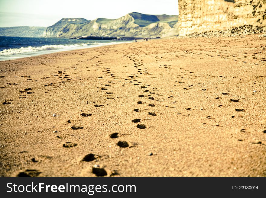 Cross process image showing trail of footsteps on a sandy beach with ocean and cliffs in background. Cross process image showing trail of footsteps on a sandy beach with ocean and cliffs in background