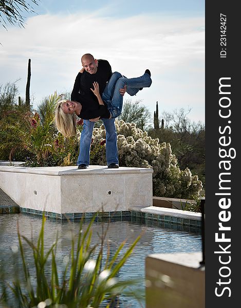 Full length of a happy playful couple posing next to a pool. He appears to be about to drop her in the pool. A green winter desert landscape is in the background. Full length of a happy playful couple posing next to a pool. He appears to be about to drop her in the pool. A green winter desert landscape is in the background.