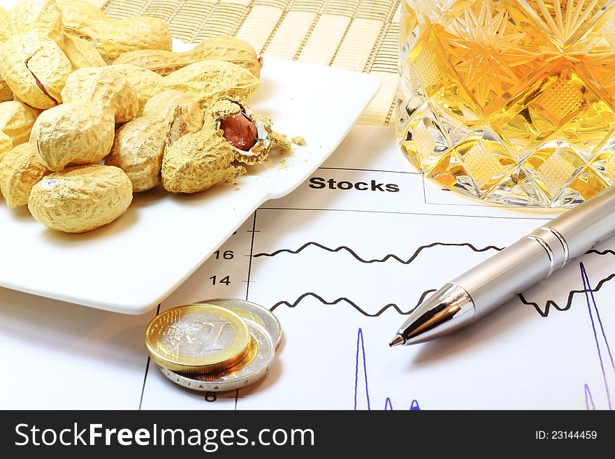 A concept of stock market, crisis and gambling with money; peanuts, coins, drink, a pen and stock graph on the table. A concept of stock market, crisis and gambling with money; peanuts, coins, drink, a pen and stock graph on the table.