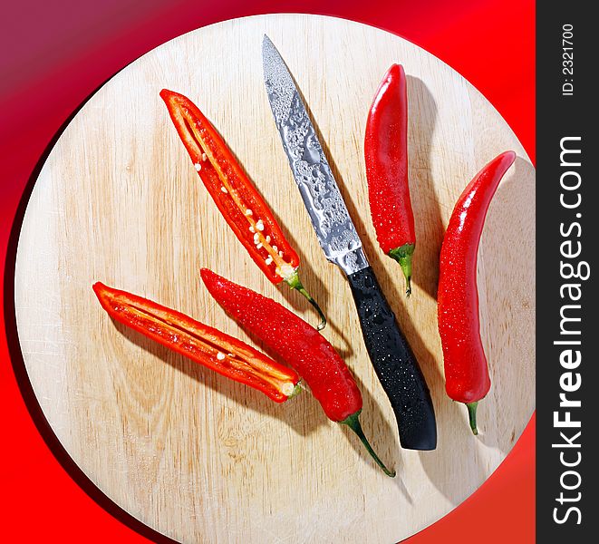 Red chili peppers and a knife, all covered with water-drops, on a preparation board. A view from above. Red background. Red chili peppers and a knife, all covered with water-drops, on a preparation board. A view from above. Red background.
