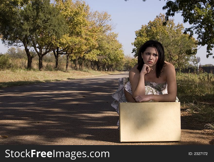 Brunette waiting roadside for a lift resting on suitcase. Brunette waiting roadside for a lift resting on suitcase