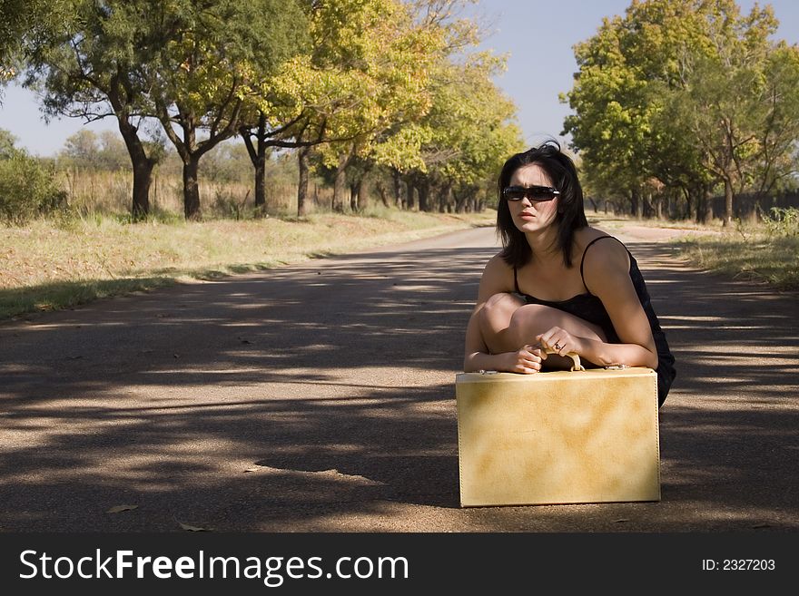 Brunette sitting next to road waiting for a lift wearing her sunglasses and holding her suitcase. Brunette sitting next to road waiting for a lift wearing her sunglasses and holding her suitcase