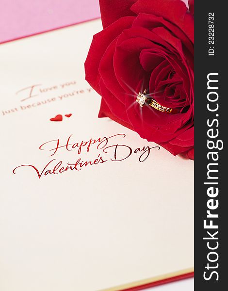 Photo of A gold and diamond ring embedded on a red rose and a valentines day greeting card with a pink background. Photo of A gold and diamond ring embedded on a red rose and a valentines day greeting card with a pink background.