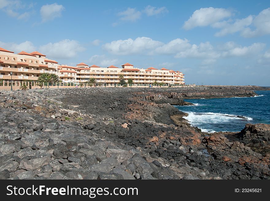 Image shows a hotel at the coast of Fuerteventura. Stone coast is black, ocean and sky are blue, sky is light cloudy. Image shows a hotel at the coast of Fuerteventura. Stone coast is black, ocean and sky are blue, sky is light cloudy.