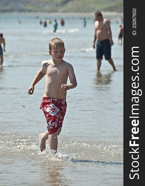 An image of a young boy wearing boardshorts running through the water on a hot summers day. An image of a young boy wearing boardshorts running through the water on a hot summers day