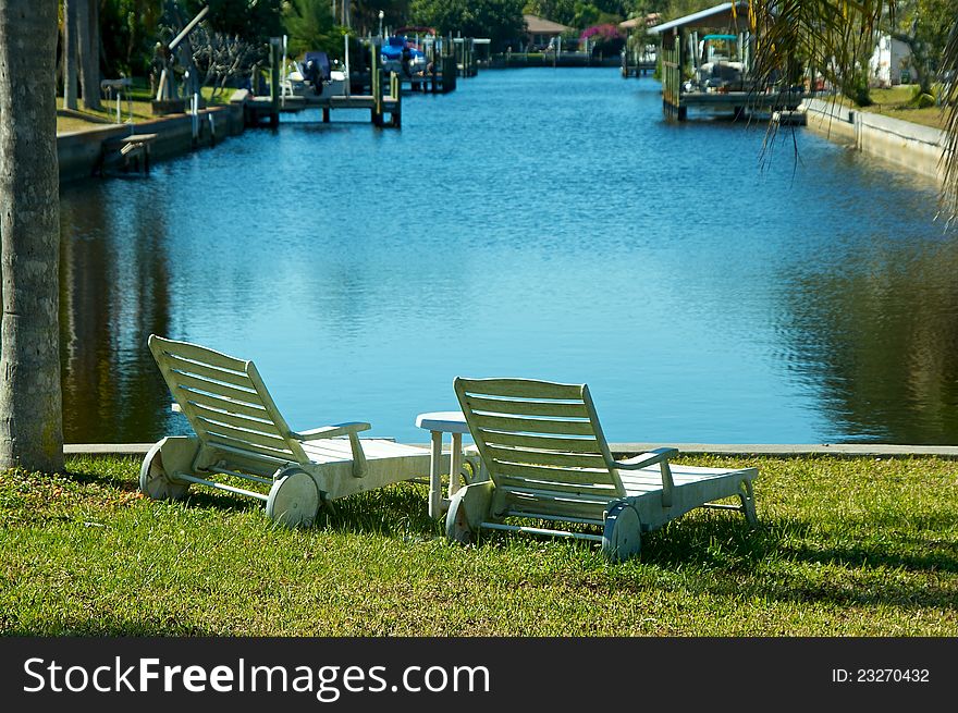 Two white outside lounge chairs are sitting by the water at the mouth of a canal in florida. Two white outside lounge chairs are sitting by the water at the mouth of a canal in florida.