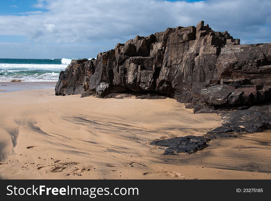 Beach of Fuerteventura, Canaria. Black stones on the right, sand, blue ocean and cloudy light blue sky above. Beach of Fuerteventura, Canaria. Black stones on the right, sand, blue ocean and cloudy light blue sky above.