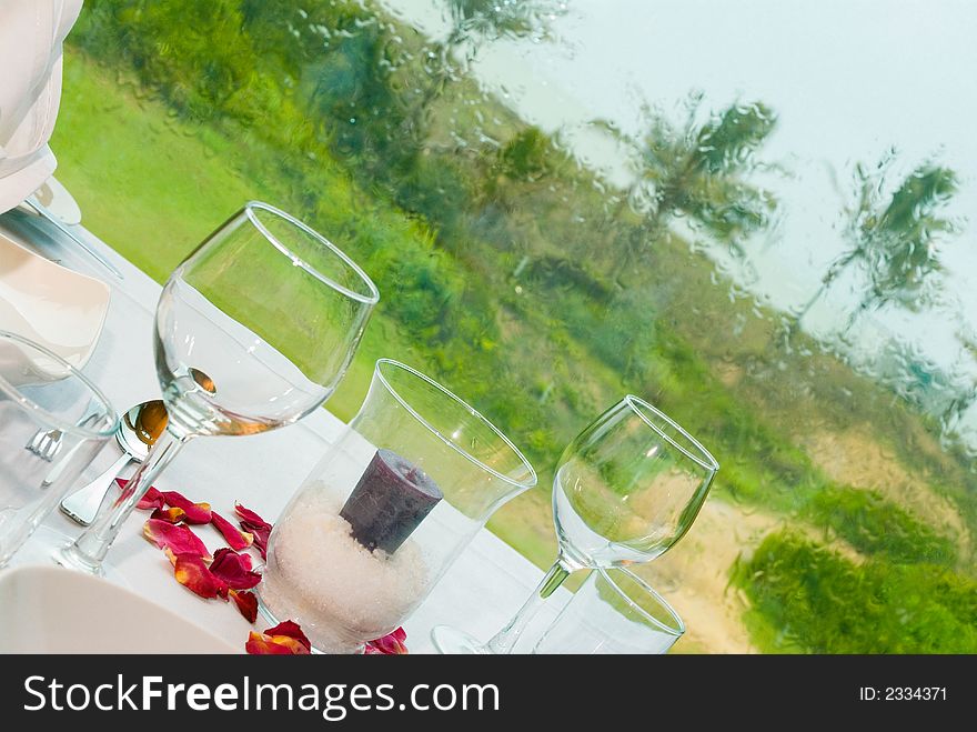 A formal dining setting with candle and rose petals as accessories up against a glass window looking out over a garden setting on a very rainy day