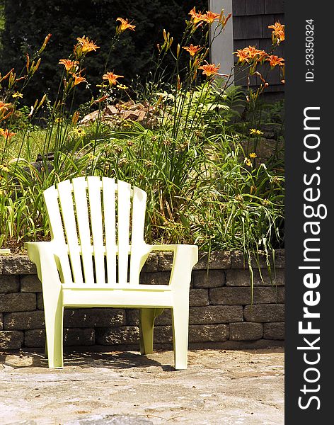 A white garden chair stands on a patio in front of a stone wall and tiger lilies. A white garden chair stands on a patio in front of a stone wall and tiger lilies