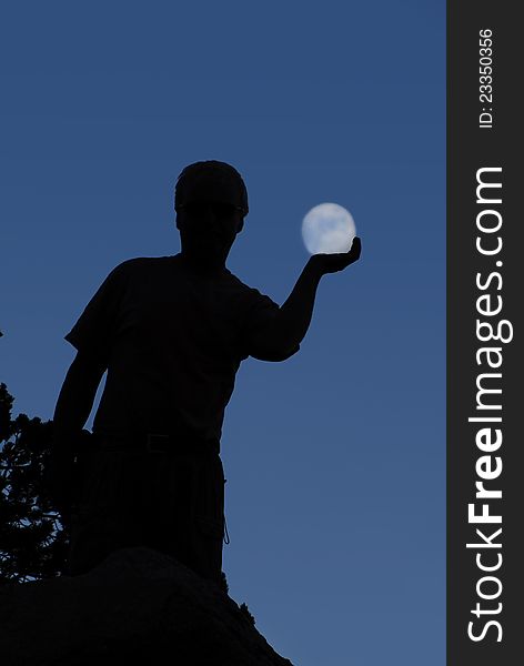 A man stands silhouetted against a dark sky with the appearance of the moon in his hand. A man stands silhouetted against a dark sky with the appearance of the moon in his hand