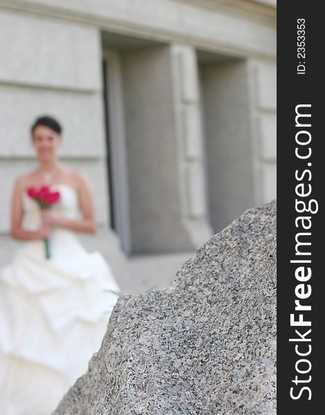 Bride Holding her bouquet standing in front of a stone building with the focus on the foreground of a large stone. Bride Holding her bouquet standing in front of a stone building with the focus on the foreground of a large stone