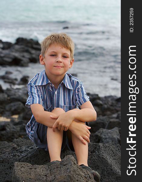 Little boy dreaming near the ocean, sitting on lava stones