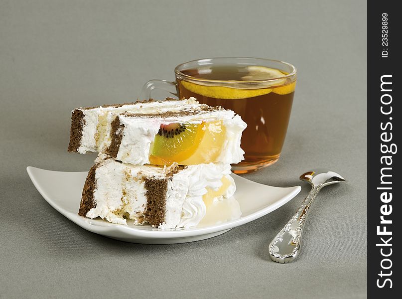 Piece of cake with fruit, tea with a lemon, tea-spoon on grey fabric. Piece of cake with fruit, tea with a lemon, tea-spoon on grey fabric