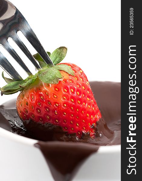 A ripe red strawberry on the end of a fork is being dipped into a bowl of melted brown chocolate. A ripe red strawberry on the end of a fork is being dipped into a bowl of melted brown chocolate