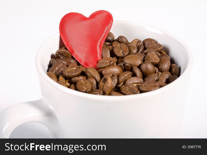 White Cup filled with coffeebeans on white background. Red heart on the beans. White Cup filled with coffeebeans on white background. Red heart on the beans.