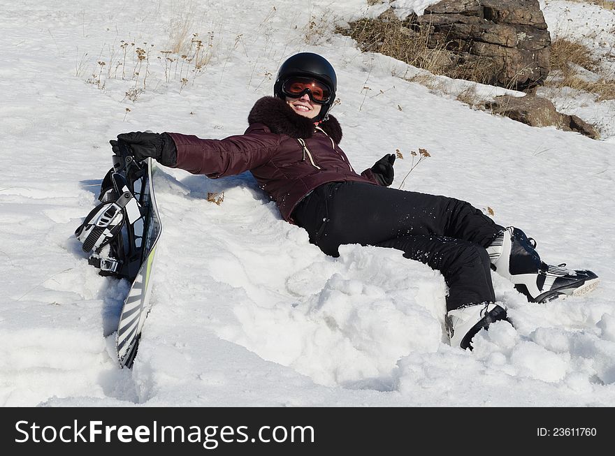 The smiling young woman in a mountain-skiing helmet, in ski glasses, lies on snow and holds a snowboard. The smiling young woman in a mountain-skiing helmet, in ski glasses, lies on snow and holds a snowboard