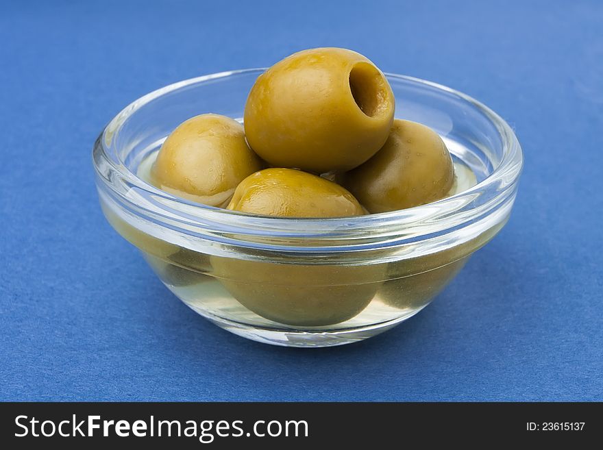 Green olives in a transparent bowl against blue background. Green olives in a transparent bowl against blue background