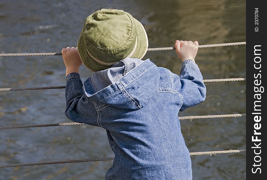 Child with denim hat and jacket looking through a fence onto water. Child with denim hat and jacket looking through a fence onto water