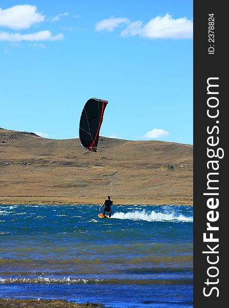 Black and red kitesurfer cruising on water at scenic dam with mountain behind, blue sky and fress water
