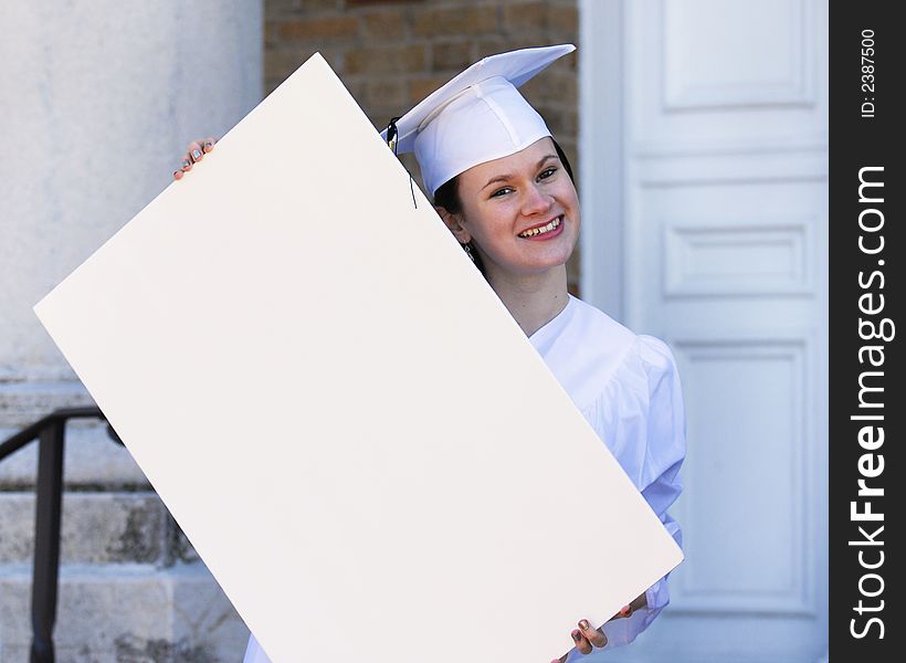 Beautiful graduate in white cap and gown holding a blank sign in front of her school. Beautiful graduate in white cap and gown holding a blank sign in front of her school.