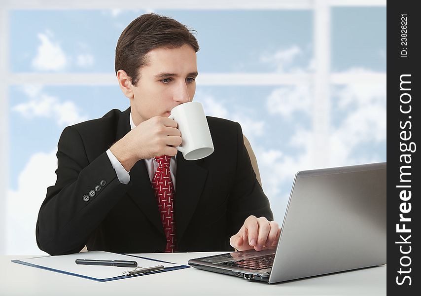 Young successful business man working behind the notebook, sitting at a desk with a cup of coffee on the background of a window to the sky and clouds. Young successful business man working behind the notebook, sitting at a desk with a cup of coffee on the background of a window to the sky and clouds