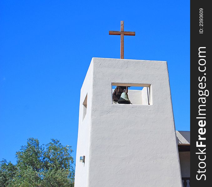 White stucco church bell tower with wooden cross atop against a blue sky