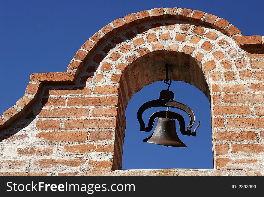 An iron bell in a rustic, red brick arch against a clear blue sky in the American Southwest. An iron bell in a rustic, red brick arch against a clear blue sky in the American Southwest.