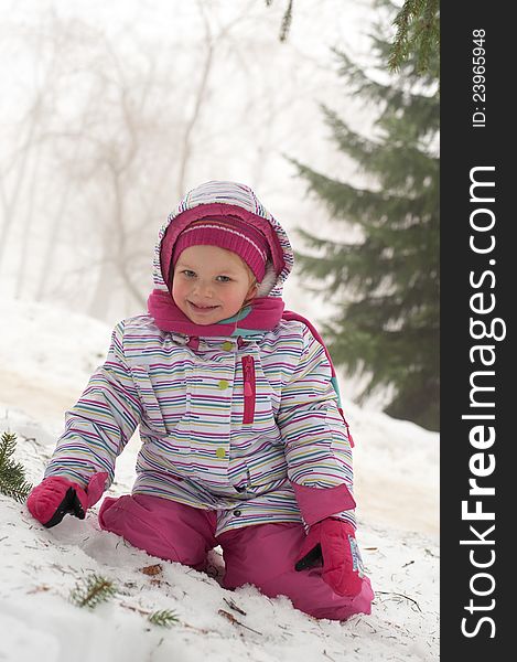 Girl sitting on the snow in the park with trees in the background, dressed in winter snow suite. Girl sitting on the snow in the park with trees in the background, dressed in winter snow suite