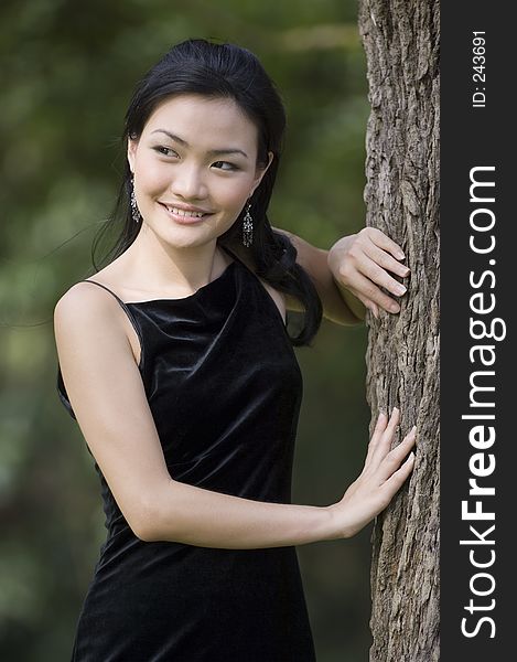 A beautiful young asian woman poses in a black evening dress next to a tree. A beautiful young asian woman poses in a black evening dress next to a tree