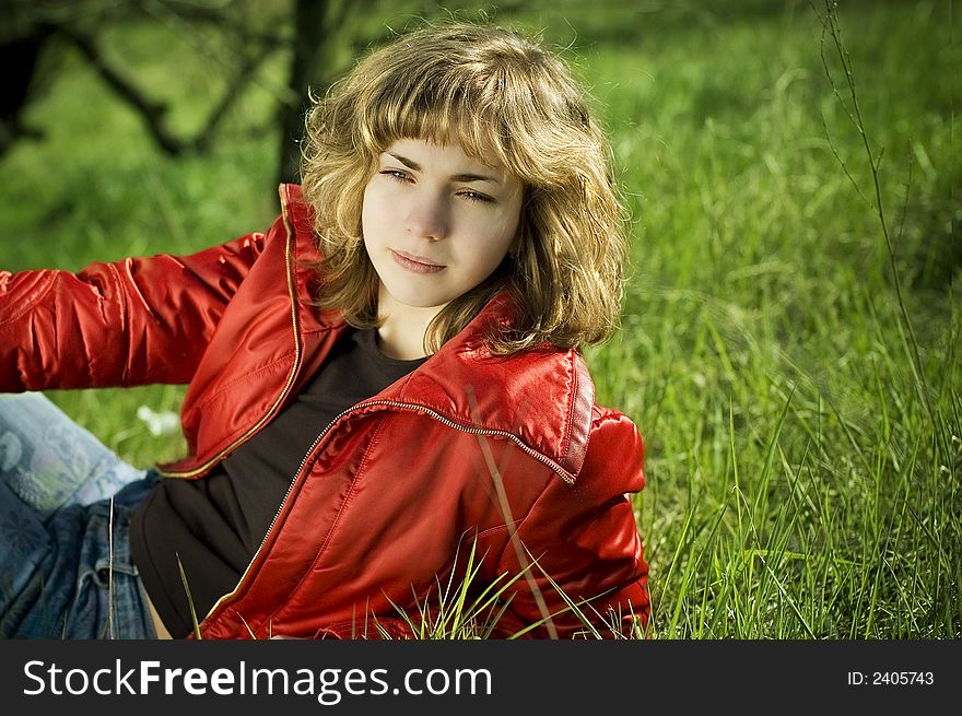 Young pretty blond wearing red jacket sitting on the green grass. Young pretty blond wearing red jacket sitting on the green grass