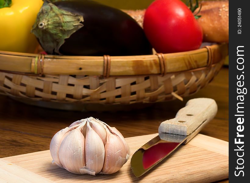 A bulb of garlic on a cutting board with knife and basket of fresh vegetables in the background