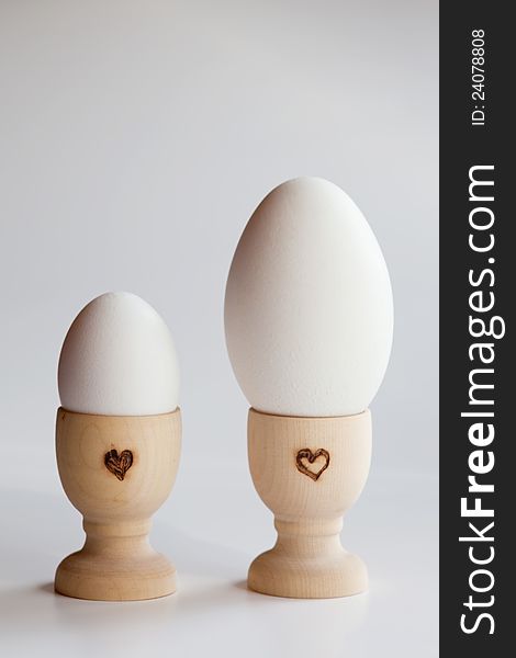 One goose egg and one hen's egg, both in wooden egg cups with a heart shape. One goose egg and one hen's egg, both in wooden egg cups with a heart shape