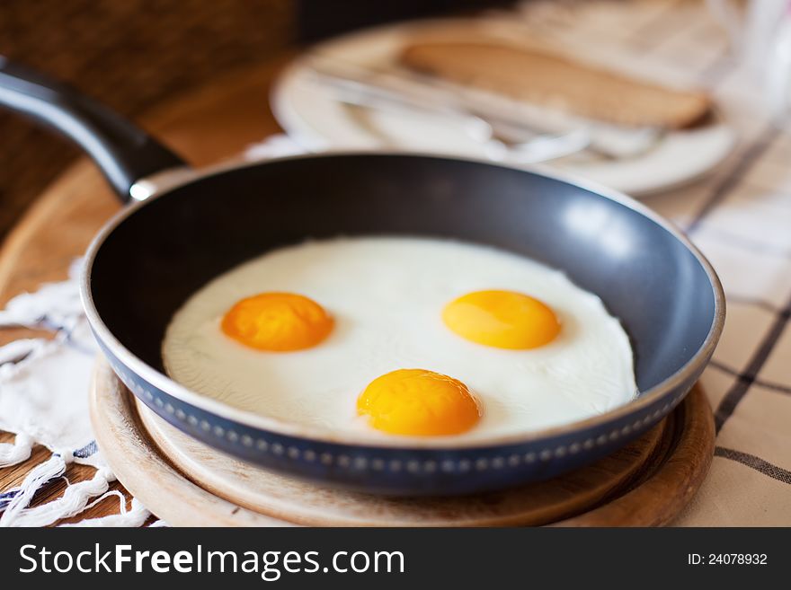 Frying pan with three fried eggs on a kitchen table with a plate with cutlery and bread