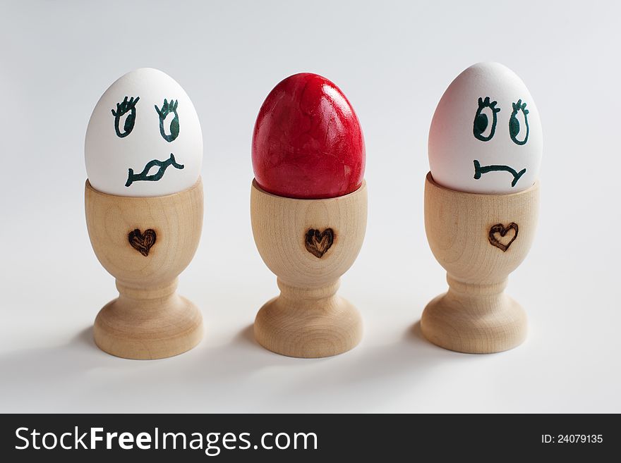 Three eggs in a egg cup, standing in a row. The egg in the middle is a red colored easter egg and the left and right one are looking surprised two the easter egg. Three eggs in a egg cup, standing in a row. The egg in the middle is a red colored easter egg and the left and right one are looking surprised two the easter egg.