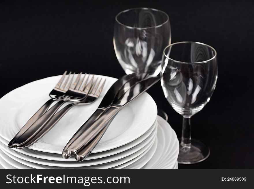 Two wine glasses, a stack of white plates, knives and forks on a black background. Two wine glasses, a stack of white plates, knives and forks on a black background