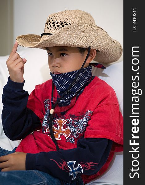 Young asian boy sitting on a white sofa wearing a cowboy hat and a bandana with finger on the brim of the hat. Young asian boy sitting on a white sofa wearing a cowboy hat and a bandana with finger on the brim of the hat